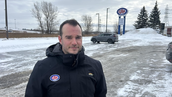 Un homme vêtu d'un manteau de CAA-Québec pose dans un stationnement devant une enseigne de CAA-Québec.