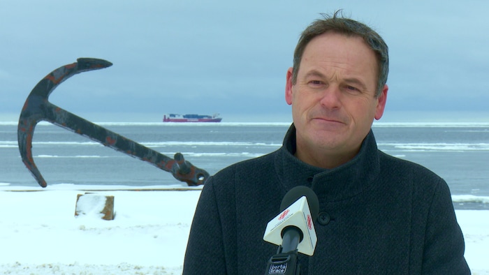 Le maire de Sainte-Anne-des-Monts, Simon Deschênes. Derrière lui, une ancre de bateau, le fleuve et un navire de marchandise au large.