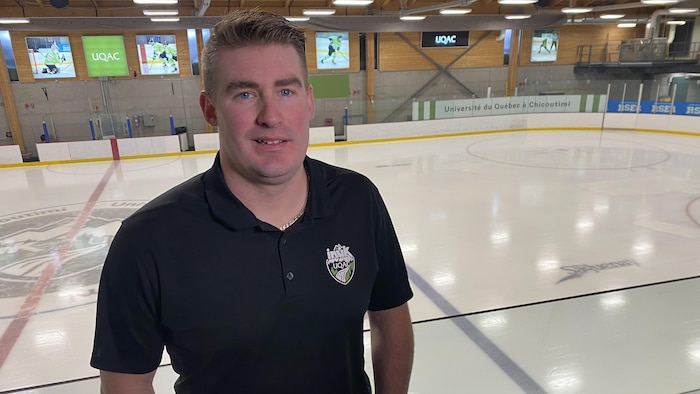 Un homme en polo noir portant le logo des Inuk de l'UQAC pose devant une patinoire intérieure. L'arrière-plan montre la glace vide, les bandes et des bannières de l'Université du Québec à Chicoutimi.