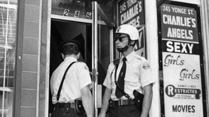 Une photo en noir et blanc montre des policiers devant un immeuble. Des affiches annoncent des spectacles pour adultes. 