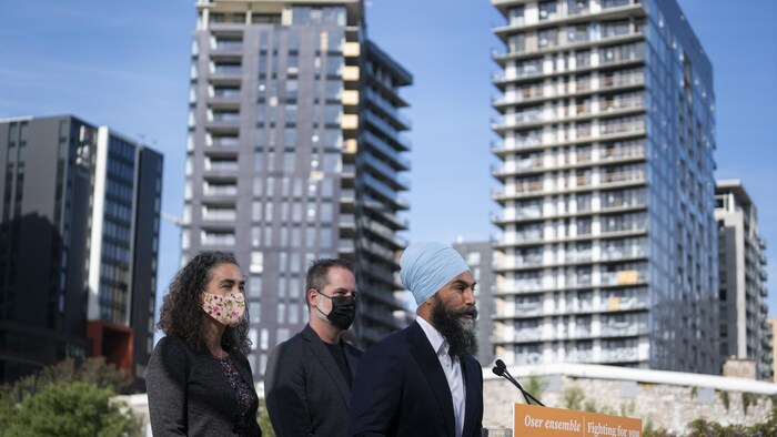 M. Singh parle aux médias, debout, de profil. Derrière lui se tiennent Mme Thiébaut et M. Boulerice. En arrière-plan, des immeubles à logements du centre-ville de Montréal.