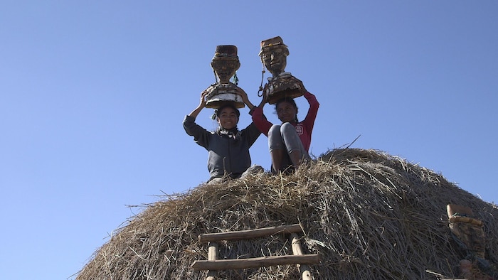 Deux fillettes sont accroupies sur le toit d'une hutte. Elles tiennent une statuette sur leur tête.
