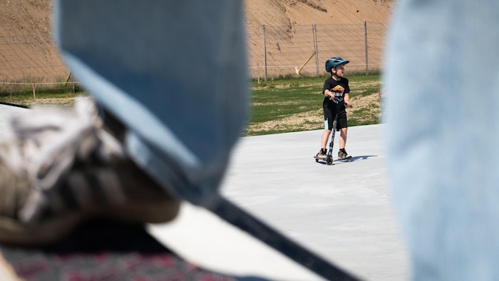 Un enfant sur une trottinette, avec, en avant plan les pieds de quelqu'un sur une planche à roulette.