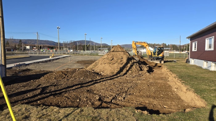 Une pelleteuse creuse un sol, entre un bâtiment et un parking.