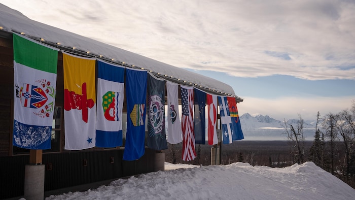 Les huit drapeaux des délégations suspendus les uns à côté des autres, en fond une chaîne de montagnes, aux Jeux d'hiver de l'Arctique 2024, à Mat-Su en Alaska. 