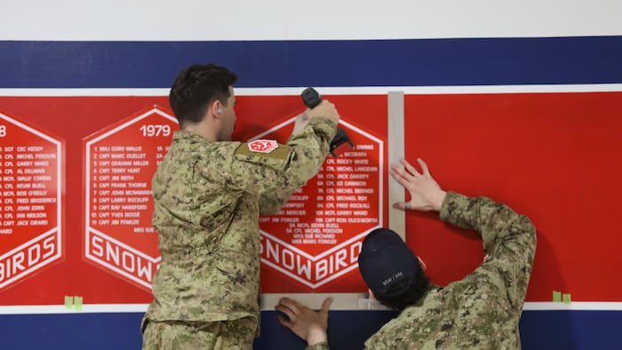Deux membres du personnel vêtus de tenues de camouflage accrochent des fanions sur lesquels figurent les noms des pilotes ayant fait partie des Snowbirds au fil des ans.