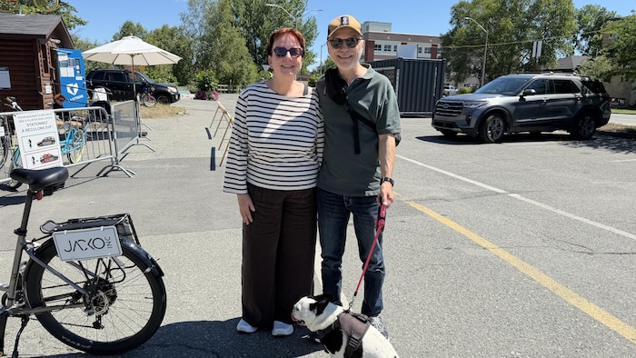 Une femme et un homme accompagnés d'un chien sourient à la caméra en posant près d'un vélo à côté d'une piste cyclable.