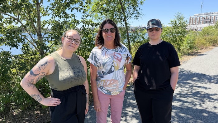 Trois femmes prennent la pose près d'un lac, sur une piste cyclable, à l'ombre de quelques arbres.