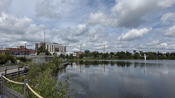 Au loin, l'hôpital de Rouyn-Noranda et les deux cheminées de la Fonderie Horne. À l'avant-plan, le lac Osisko sous un ciel nuageux.