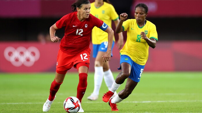 لاعبة كرة القدم الكنديّة كريستين سانكلير تركل الكرة وإلى جانبها لاعبتان من فريق البرازيل.