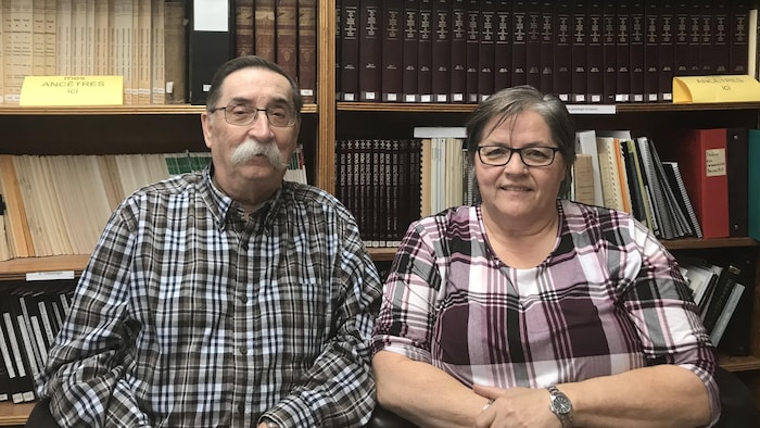 Un homme portant la moustache et une femme sourient à la caméra devant une bibliothèque remplie de livres d'époque.