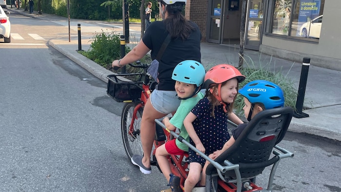 Trois enfants sont assis sur un vélo-cargo et sourient; une femme conduit et pédale.