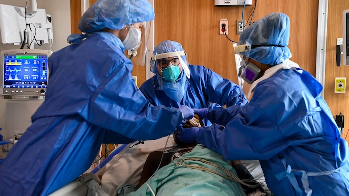 Trois infirmières en combinaisons, masques et visières s'affairent autour d'un patient alité. 