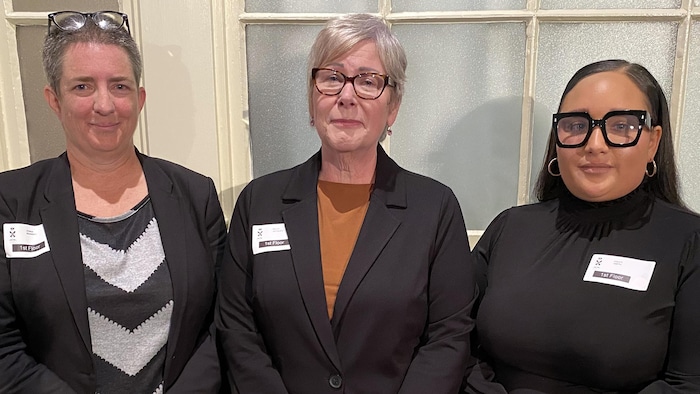 Trois femmes d'âges divers se tiennent droites, les mains jointes en tenant des documents.