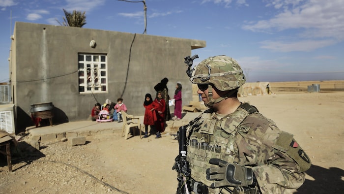 Un soldat marche devant une maison où se trouvent femmes et enfants.