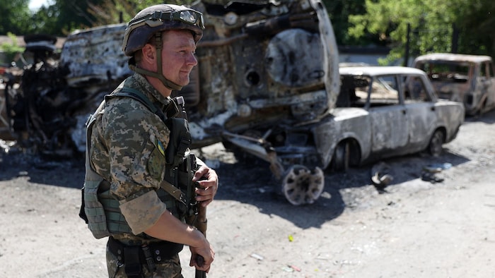 Un militaire armé marche près de carcasses de véhicules calcinées.