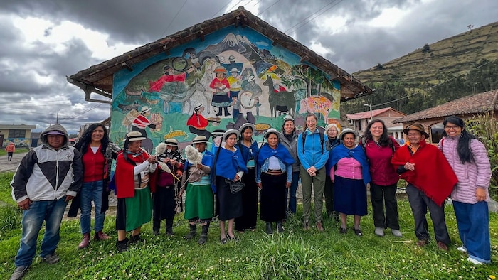 Un groupe hétérogène de personnes pose devant une maison décorée d'une fresque murale colorée représentant la vie rurale andine. On y distingue des vêtements traditionnels, comme des ponchos et chapeaux, sous un ciel nuageux en zone montagneuse.