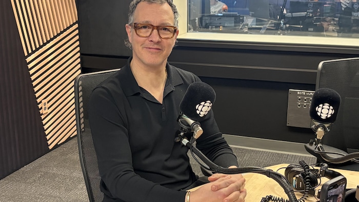 Un homme prend la pose dans un studio de radio devant un micro arborant le logo de Radio-Canada.