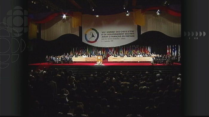 Une salle comble devant une scène sur laquelle sont assis des dirigeants des pays de la francophonie internationale et les drapeaux représentant chacun de ces pays. 