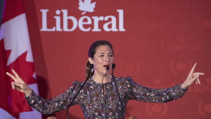 Sophie Grégoire Trudeau lève les bras en parlant au micro devant un drapeau du Canada et un logo du Parti libéral.