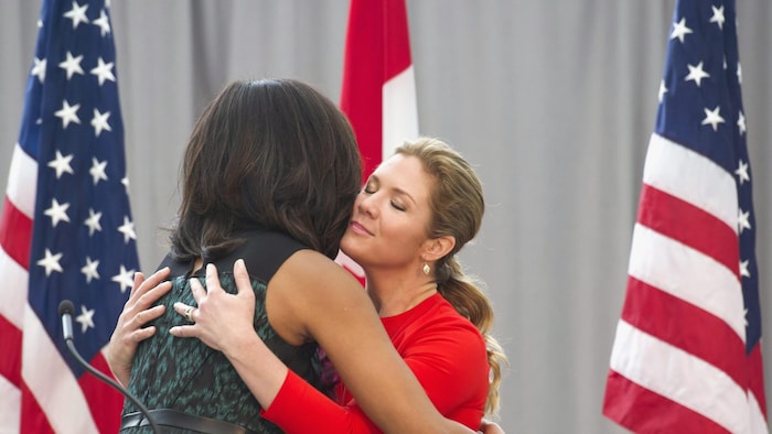 Michelle Obama et Sophie Grégoire-Trudeau s'enlacent devant des drapeaux des États-Unis et du Canada. 