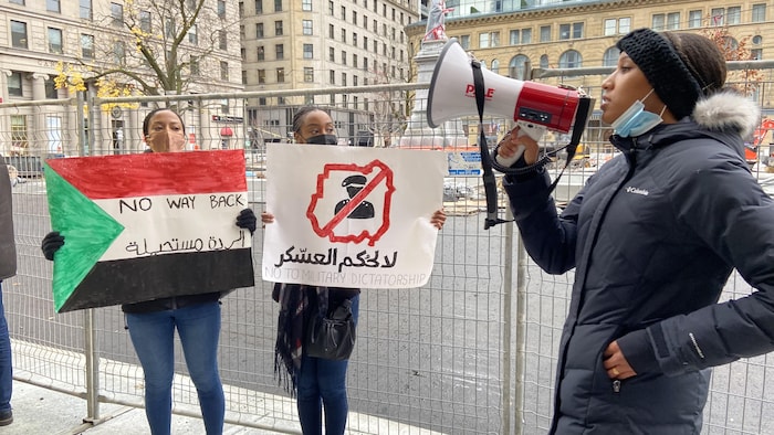 Deux jeunes filles, portant l'une un drapeau soudanais et l'autre une pancarte, et une femme qui utilise un porte-voix.
