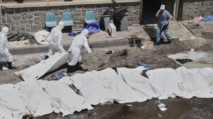 Trois travailleurs vêtus d'habits de protection s'affairent autour d'une dizaine de corps alignés dans une rue, couverts de draps blancs.