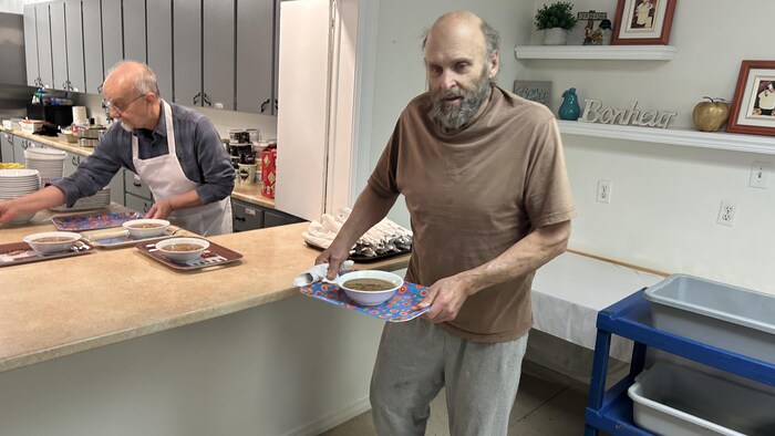 Un homme marche dans un local, un cabaret à la main sur lequel il y a une soupe. Derrière lui, une personne vêtue d'un tablier préparent d'autres soupes dans une cuisine.