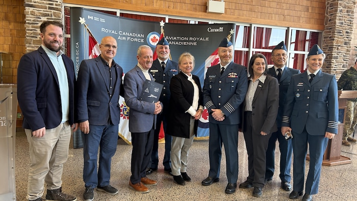 Neuf personnes, dont certaines en uniforme, posent en souriant devant des drapeaux canadiens et une bannière de l'aviation royale canadienne.