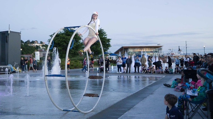 Une femme dans une grande structure en forme de double anneau en équilibre sur une place publique devant des spectateurs.