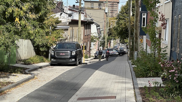 Une voiture de police barre l'accès à une rue résidentielle en pente où marchent deux agents.