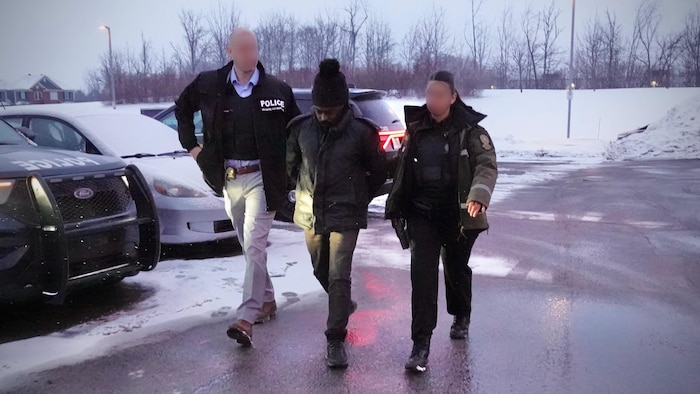 Deux policiers, dont une femme, encadrent un suspect menotté dans un stationnement en hiver.
