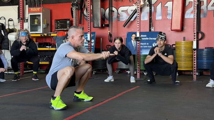 Un entraîneur aux chaussures vertes effectue une flexion des jambes (squat), bras tendus, dans une salle d'entraînement.
