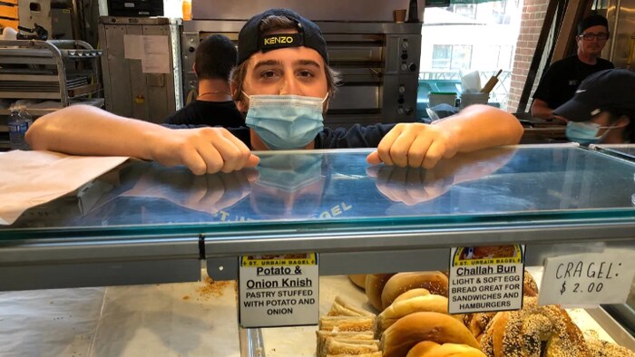 Un jeune homme portant un masque et une casquette, les deux mains sur son comptoir, derrière la devanture de sa boutique de bagel.
