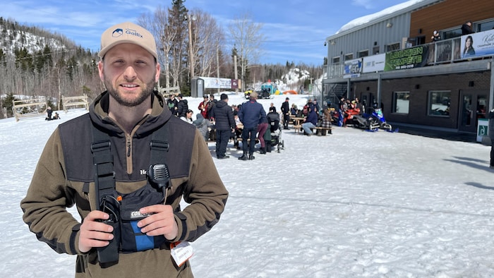 Un homme sourit à la caméra, en avant-plan d'un bâtiment et de plusieurs dizaines de personnes rassemblées autour de tables à l'extérieur en hiver.