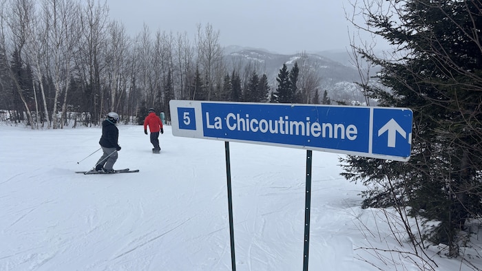 Au premier plan, un panneau bleu indique la piste « La Chicoutimienne » avec le chiffre 5. À l'arrière-plan, deux skieurs descendent une pente enneigée bordée d'arbres dépouillés, sous un ciel couvert de montagne.