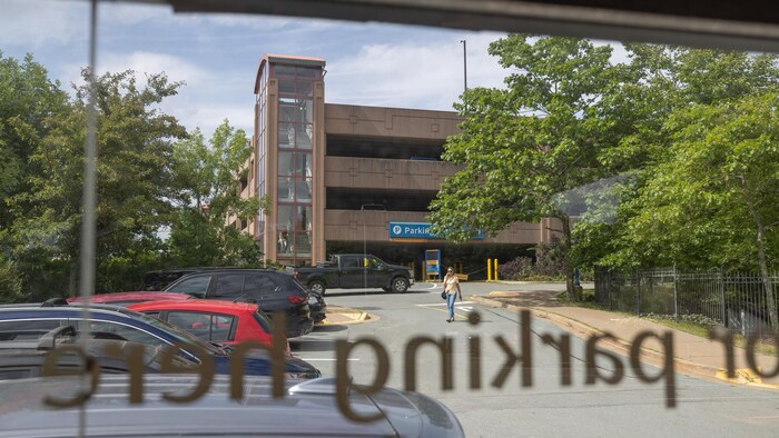 À travers une porte sur laquelle le mot «Parking» apparaît écrit à l'envers, on voit une femme qui marche à l'extérieur en parlant au téléphone devant un stationnement à étages en béton.