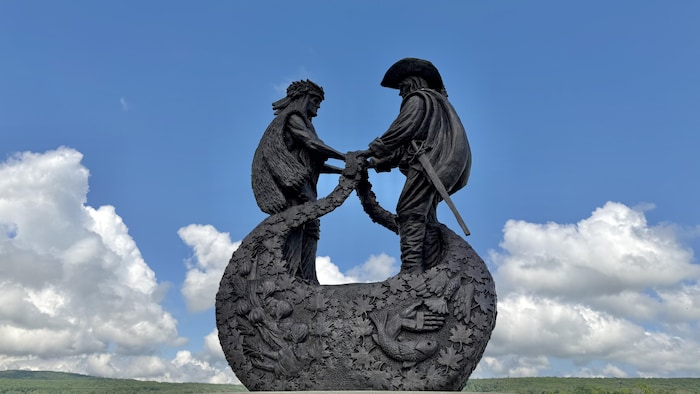 Sculpture de bronze sombre représentant l'explorateur Samuel de Champlain et un chef autochtone wendat debout face à face, sur un pied d'égalité, se tenant la main au-dessus d'une structure en demi-lune ornée de motifs sculptés.