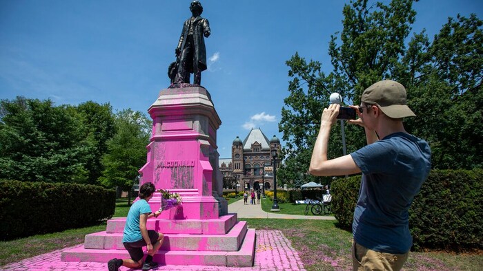 Le socle de la statue est recouvert de peinture rose. Une femme dépose une gerbe de fleurs à ses pieds tandis qu'un homme prend la scène en photo.