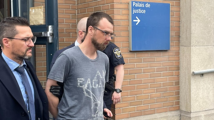 Un homme menotté les mains dans le dos sort d'un bâtiment escorté par deux hommes en civil et des policiers.