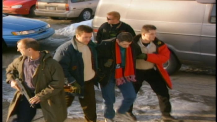 Un homme escorté par la Sûreté du Québec avec des policiers armés jusqu'aux dents. 