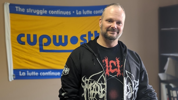 Stéphane Genest devant le logo du Syndicat des travailleurs et travailleuses des postes.
