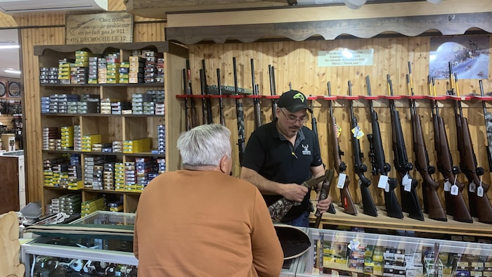 Deux hommes debout dans un commerce. Des fusils de chasse et des munitions derrière le comptoir.