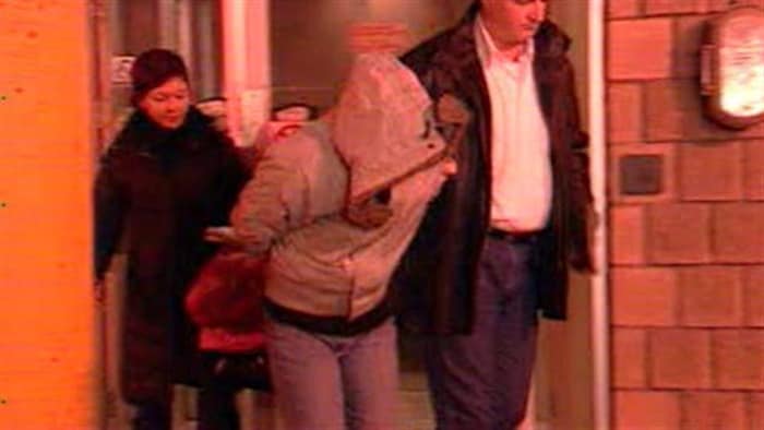 Une femme sort escortée par deux policiers, un capuchon sur la tête.