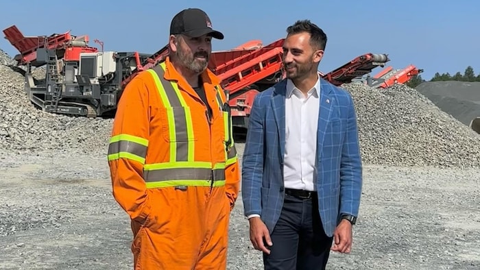 Stephen Lecce et un ouvrier marchent et parlent ensemble dans un champ de gravier, devant de la machinerie lourde.