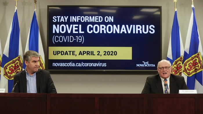Deux hommes devant un écran sur lequel apparaît le mot coronavirus et la date du jour.