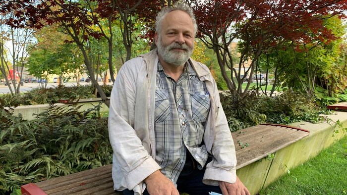 A man is sitting on a bench in a park, in front of various plants and small trees.