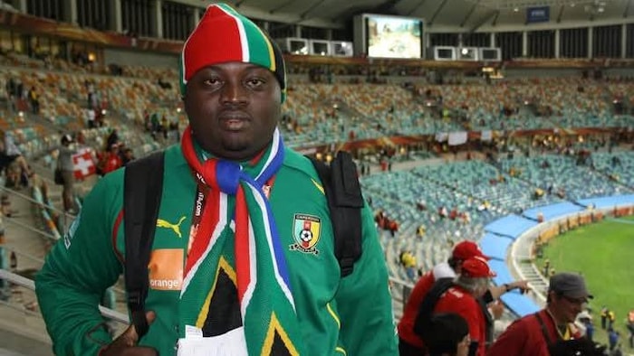 Dans les gradins d’un stade, Steve Djouguela porte une veste aux couleurs des Lions indomptables du Cameroun.