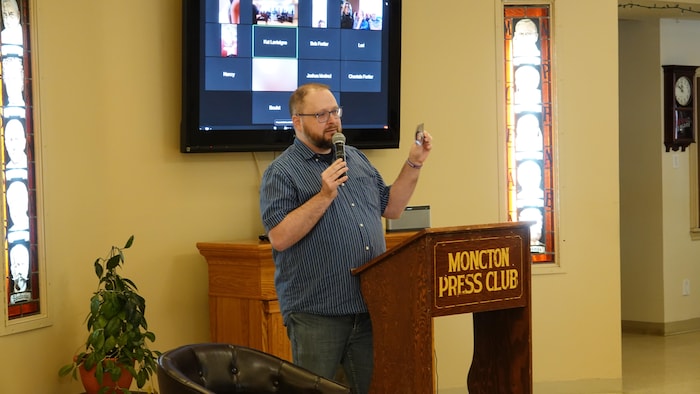 Un homme montre une photo. Il est derrière un lutrin et semble s'adresser à une foule.