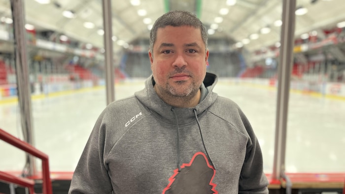 Steve Hartley porte un gilet avec une tête de chien qui aboie, le logo des Huskies. Il se tient debout devant la patinoire de l'équipe de hockey.